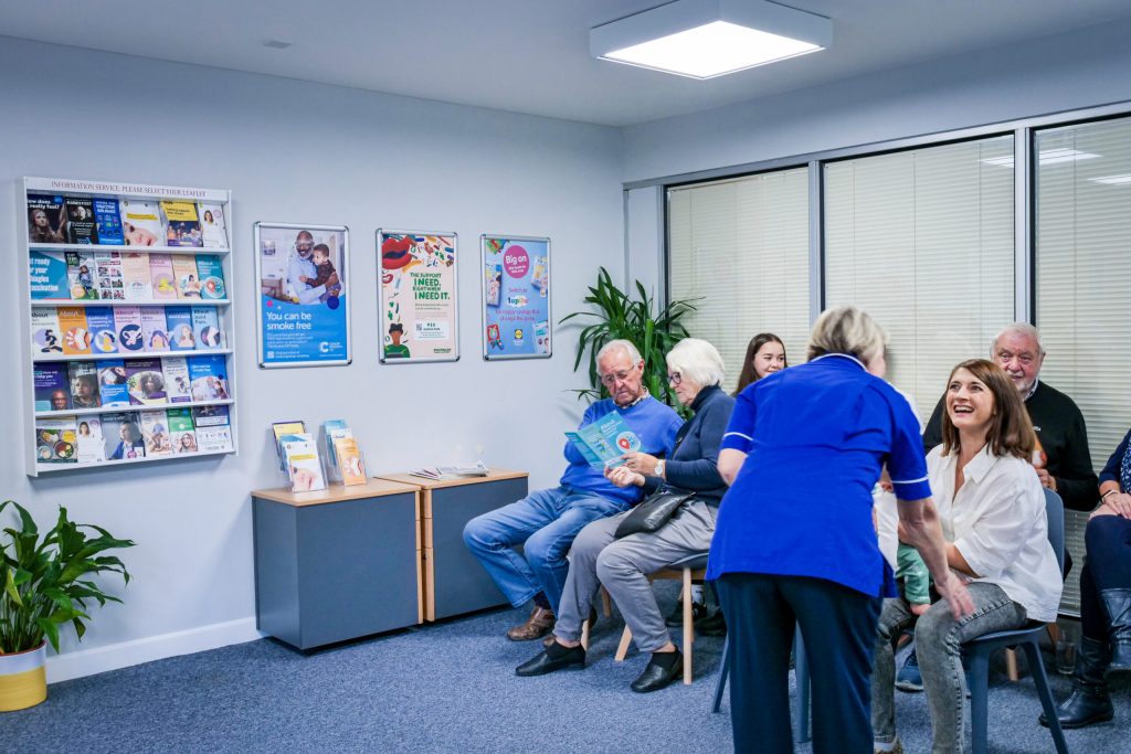 People sit on two rows of plastic chairs in a brightly lit medical waiting room with informational posters on the wall, and a nurse wearing blue scrubs leaning over one waiting woman.