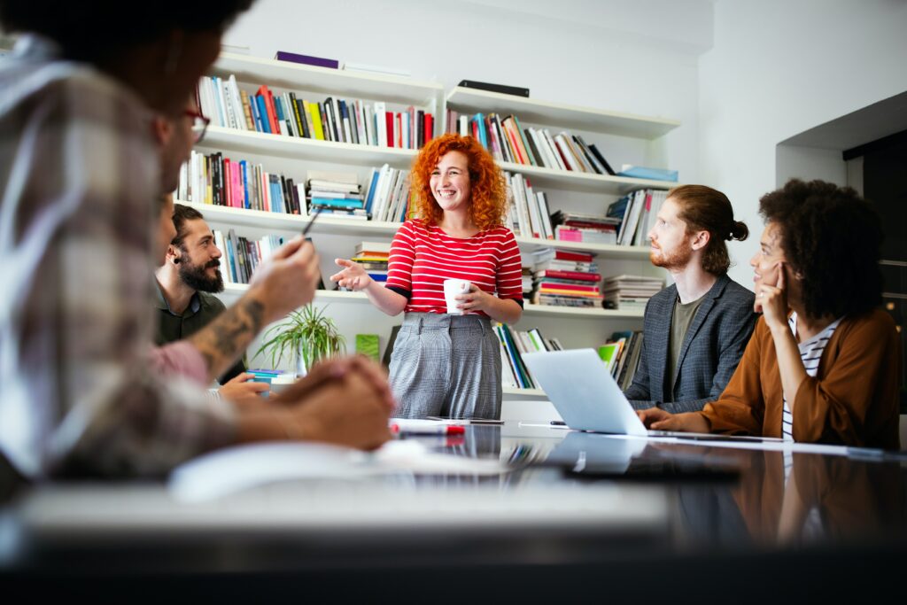 A white woman with curly ginger hair wearing a stripey red and white top speaks with others around a desk, laughing and smiling