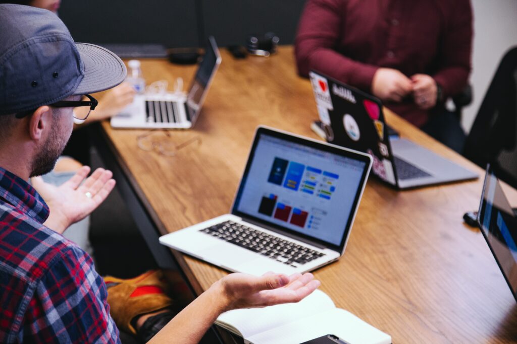 A white man wearing a cap and plaid shirt sits at a desk with other people, he is shrugging, has his back to the camera and has a laptop