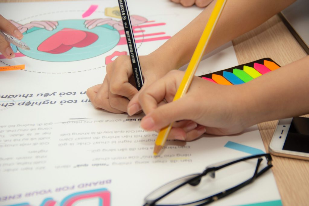 A close-up of three childrens' hands drawing on a print-out with pens and coloured pencils.