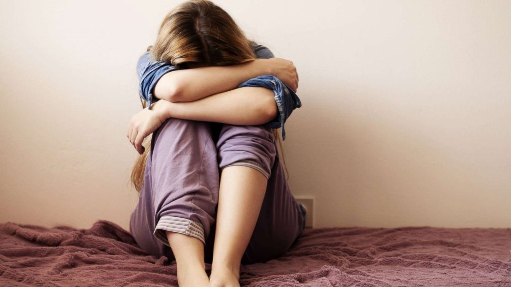 Young woman sitting on a bed with her head resting on her knees and arms wrapped around her legs, appearing distressed or overwhelmed.